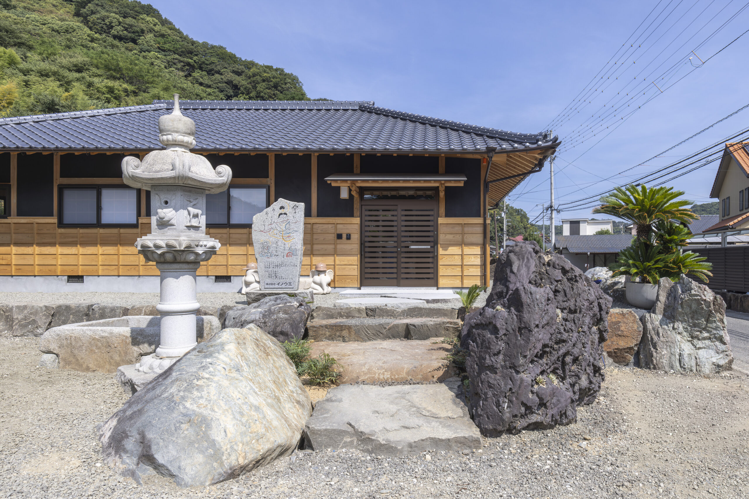瓦屋根の平屋の和風住宅外観で、木の格子の腰壁と濃茶の引き戸玄関があり、手前には石灯籠や大きな庭石、石の飛び石が配されている。